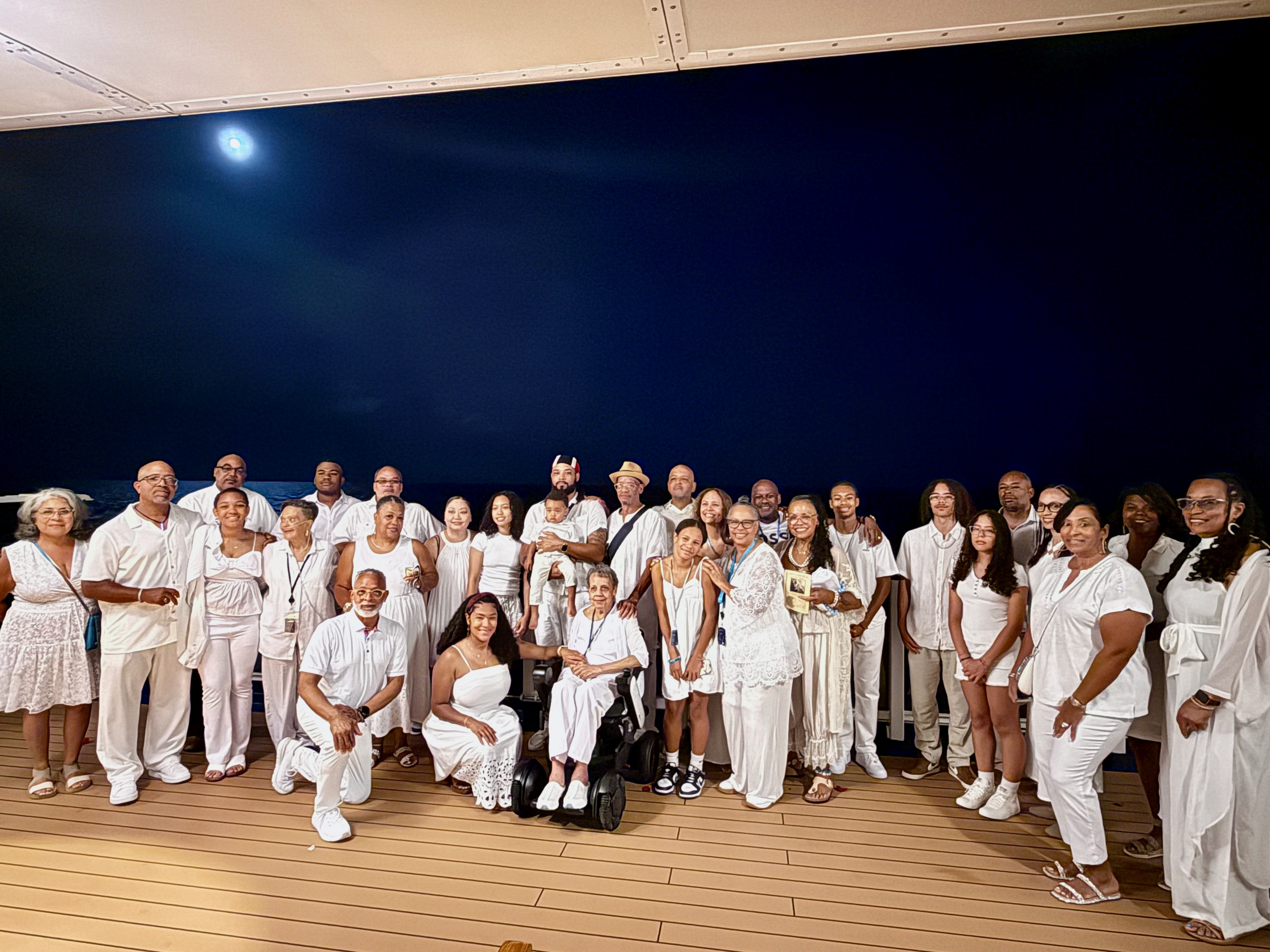 A large family gathering all dressed in white, posing on a deck at sea under the night sky