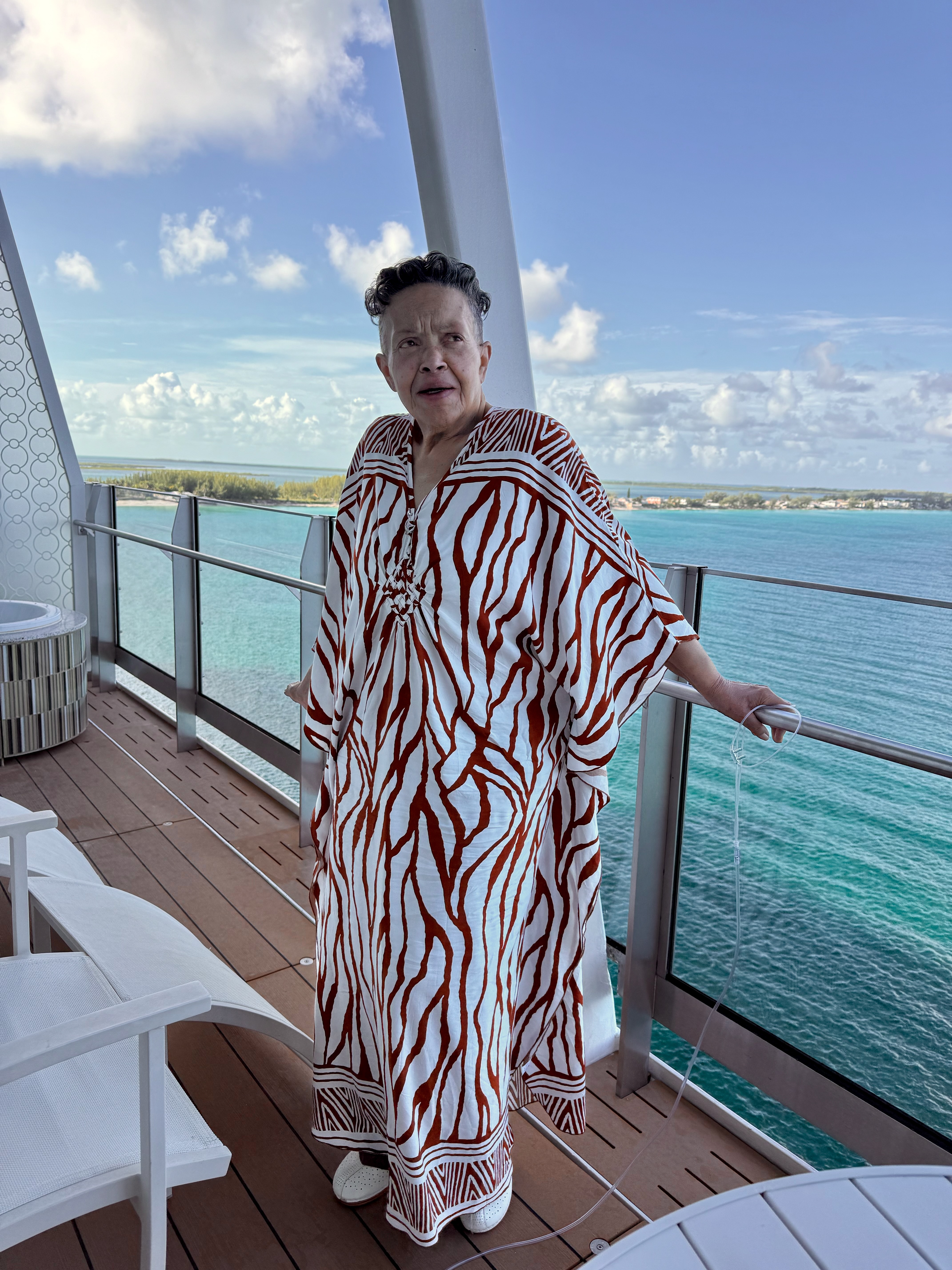 Karen on a balcony in a striking red and white caftan, the turquoise sea behind her